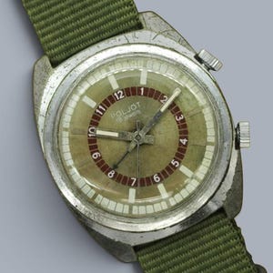 Op de afbeelding: Een vintage Poljot polshorloge met een zilverkleurige metalen kast en een groene nylon band. De ronde wijzerplaat heeft een beige centrum, een donkerrode binnenring met witte cijfers van 1 tot 12 en witte uurmarkeringen. Twee kronen aan de zijkant.
