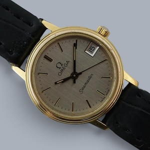 May include: An Omega Seamaster wristwatch featuring a round gold-tone case, a textured champagne-colored dial with black hour markers, a date window at the three o'clock position, and a black leather strap with a visible embossed crocodile pattern.