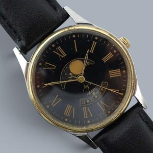Könnte beinhalten: Eine Armbanduhr mit schwarzem Lederarmband und rundem goldfarbenem Gehäuse. Das schwarze Zifferblatt zeigt goldene römische Ziffern, goldene Zeiger und eine Mondphasenanzeige oben. Ein kleines Datumsfenster befindet sich am unteren Rand.