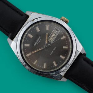 Könnte beinhalten: Eine Vintage-Cardinal-Armbanduhr mit schwarzem Lederarmband. Das Zifferblatt ist dunkel mit silbernen Akzenten und zeigt den Tag und das Datum an. Die Uhr befindet sich vor einem türkisfarbenen Hintergrund.