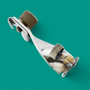 Op de afbeelding: Een zilverkleurige metalen naaivoet voor een naaimachine met een platte, schuine voetplaat, een centrale verticale steun en twee gekartelde, messingkleurige stelschroeven met schroefdraad voor bevestiging aan een naaimachine.