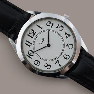 May include: A wristwatch with a round silver-tone case and a white dial featuring black Arabic numerals and the text "Luch" printed in the center. The watch has thin black hands and is attached to a black leather strap with a textured finish.