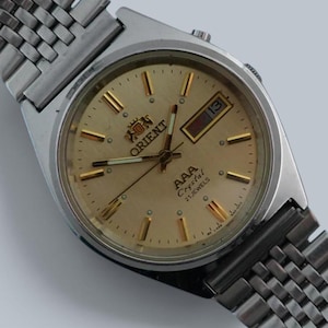 Puede incluir: Un reloj de pulsera Orient AAA Crystal 21 Jewels en tono plateado con esfera color champán, marcadores de hora dorados y una ventana de día y fecha en la posición de las tres. El reloj cuenta con una correa de metal de eslabones múltiples.