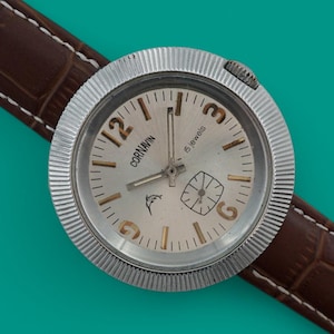 May include: A vintage wristwatch with a silver-colored face and a brown leather band. The watch face features the brand name "CORNAVIN" and a small dolphin logo. The band is a dark brown with a crocodile pattern.