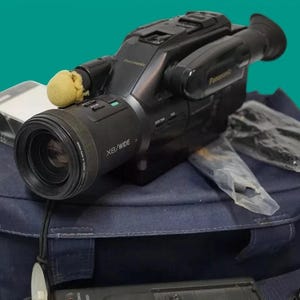 Op de afbeelding: Een zwarte Panasonic camcorder met een X8/WIDE-lens, voorzien van een kleine beige schuimrubberen hoes op de microfoon. De camcorder ligt op een donkerblauwe draagtas naast een doorzichtig cassettehoesje en een verpakt accessoire.