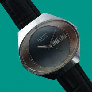 May include: A vintage wristwatch with a black face and a silver-colored case. The watch has a black leather band and displays the day and date. The watch face has the word "POLET" printed on it.