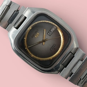 Può includere: Un orologio da polso vintage color argento con quadrante quadrato e quadrante grigio. L'orologio presenta lancette dorate e un anello interno dorato. La data indica "JUE 7". Il cinturino è composto da maglie in metallo color argento.