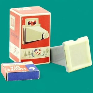 Puede incluir: Un visor de diapositivas vintage con cuerpo de plástico color crema, acompañado de su caja de cartón original roja y blanca con una ilustración gráfica del dispositivo y una pequeña caja azul y roja etiquetada como HAUFE DIA 6.