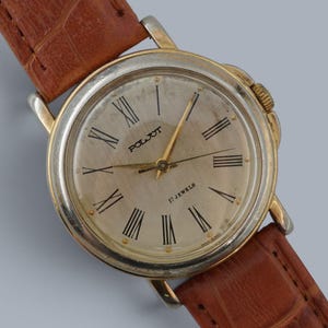 May include: A vintage Poljot wristwatch featuring a round gold-tone case, a cream-colored dial with black Roman numeral hour markers, and three thin hands. The watch is attached to a textured brown leather strap with a crocodile-embossed pattern.