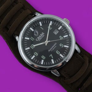 Puede incluir: Un reloj de pulsera con esfera negra y caja plateada, con números y agujas blancos. El reloj está sobre una correa de cuero marrón. La esfera del reloj dice "GJIABA Automatic" y tiene una pantalla de fecha.