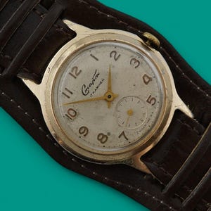 Puede incluir: Un reloj de pulsera vintage en tono dorado con esfera crema y números negros, con la marca "Gapm". El reloj está colocado en una correa de cuero marrón oscuro estilo brazalete.