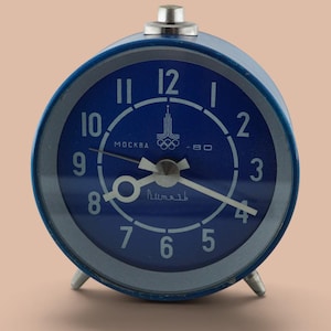 May include: A round blue analog alarm clock with a silver metal frame and two small feet. The clock face features white numerals from 1 to 12, white hands, and the text "MOCKBA-80" with an Olympic rings symbol above the center and Cyrillic text below.