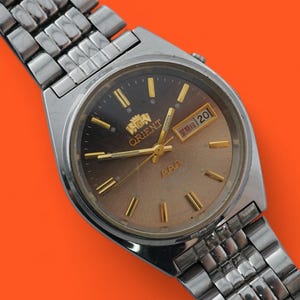 Könnte beinhalten: Eine Vintage Orient-Uhr mit silberfarbenem Metallarmband und Gehäuse. Das Zifferblatt ist ein Farbverlauf aus Braun und Gold mit goldfarbenen Zeigern und Markierungen. Das Datum wird auf 3 Uhr angezeigt. Die Uhr befindet sich vor einem orangefarbenen Hintergrund.