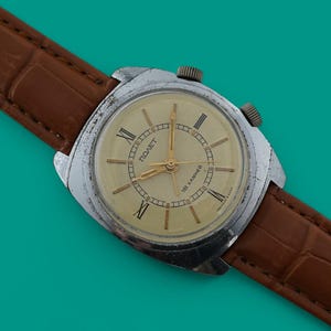 May include: A vintage wristwatch with a silver case and a cream face, displaying Roman numerals and gold hands. It has a brown leather strap with a crocodile skin effect. The watch is set against a teal background.