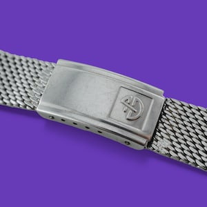 May include: A silver-toned watch band with a mesh design. The band features a rectangular clasp with a logo. The band is made of a woven metal material and is set against a purple background.