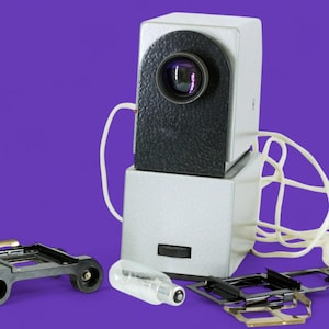 Könnte beinhalten: Ein Vintage-Diaprojektor mit silbernem und schwarzem Gehäuse, einem Objektiv und einem weißen Kabel. Der Projektor steht auf einem violetten Hintergrund. Es gibt auch zwei Diaschubladen und eine Glühbirne.