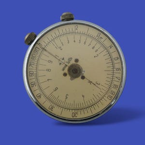 May include: A circular slide rule with a cream face and black markings, set in a silver metal frame. It features two small knobs and a series of numbers and graduations for calculations. The device is set against a blue background.