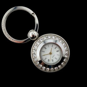 Può includere: Un portachiavi argentato con un quadrante di orologio rotondo circondato da strass. Il quadrante dell'orologio ha lancette nere e uno sfondo bianco.