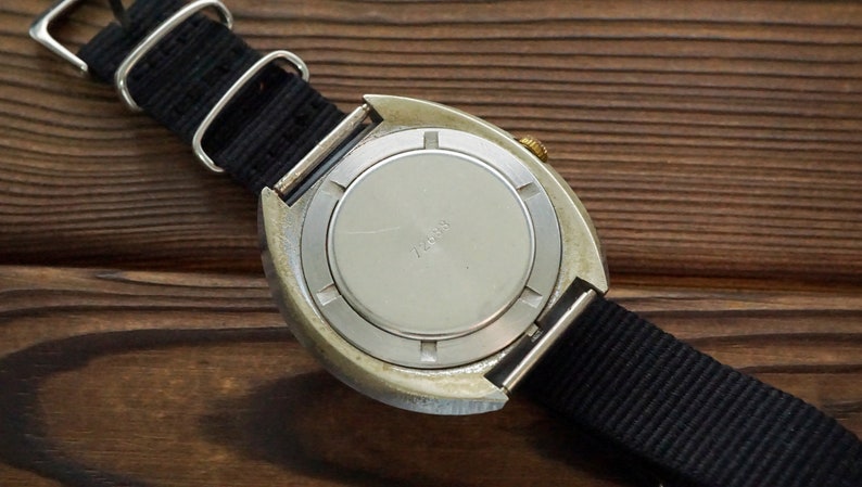 Pode incluir: A parte de tr&aacute;s de um rel&oacute;gio de pulso prateado com uma pulseira de nylon preta. O mostrador do rel&oacute;gio tem uma caixa de metal prateada com uma coroa dourada. O mostrador do rel&oacute;gio tem o texto "724018" impresso nele.