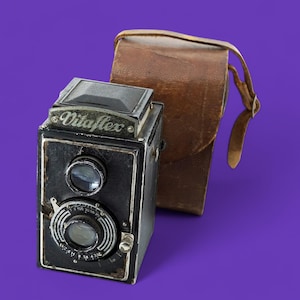 May include: A vintage black VitaFlex camera with a brown leather case. The camera has two lenses and a silver dial. The case has a strap. The camera and case are set against a purple background.