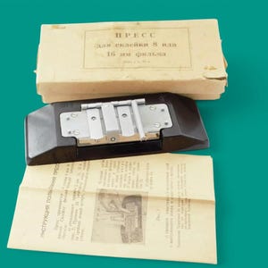 May include: A black film splicing press with a metal hinge mechanism, accompanied by a beige cardboard box labeled with the text "ПРЕСС для склейки 8 или 16 мм фильма" and a folded instruction sheet featuring technical diagrams and Russian text.