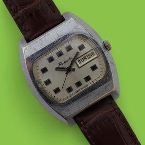 May include: Vintage Raketa watch with a silver-tone case and a brown leather strap. The watch face is off-white with black square hour markers and black hands. The day and date are displayed at the 3 o'clock position. The watch is set to Sunday the 26th.