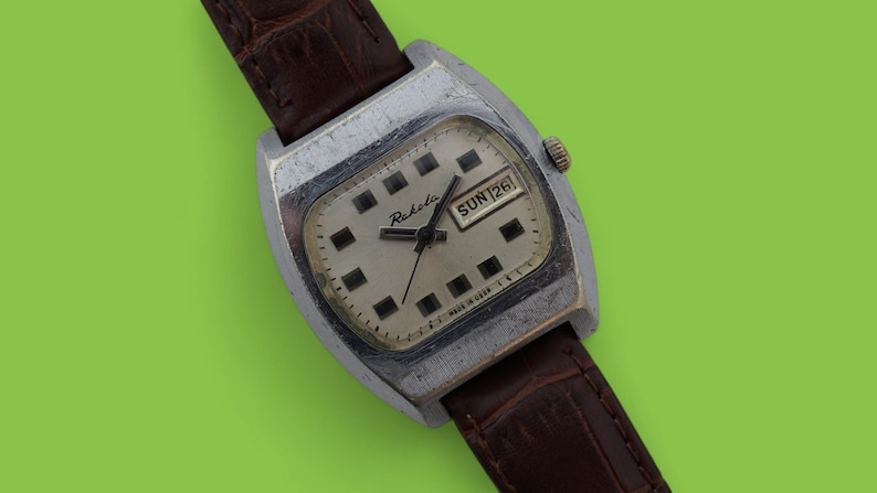 May include: Vintage Raketa watch with a silver-tone case and a brown leather strap. The watch face is off-white with black square hour markers and black hands. The day and date are displayed at the 3 o'clock position. The watch is set to Sunday the 26th.