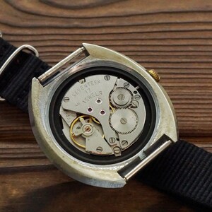 Pode incluir: A parte de tr&aacute;s de um rel&oacute;gio de pulso prateado com uma pulseira de nylon preta. O mostrador do rel&oacute;gio est&aacute; aberto, revelando o funcionamento interno do rel&oacute;gio, incluindo uma coroa de corda dourada e o texto "Seventeen Jewels".