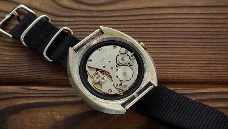 Pode incluir: A parte de tr&aacute;s de um rel&oacute;gio de pulso prateado com uma pulseira de nylon preta. O mostrador do rel&oacute;gio est&aacute; aberto, revelando o funcionamento interno do rel&oacute;gio, incluindo uma coroa de corda dourada e o texto "Seventeen Jewels".