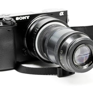 K&ouml;nnte beinhalten: Schwarze Sony Alpha Kamera mit gro&szlig;em Objektiv. Die Kamera hat einen schwarzen Riemen und befindet sich vor einem wei&szlig;en Hintergrund. Das Objektiv hat Messmarkierungen.