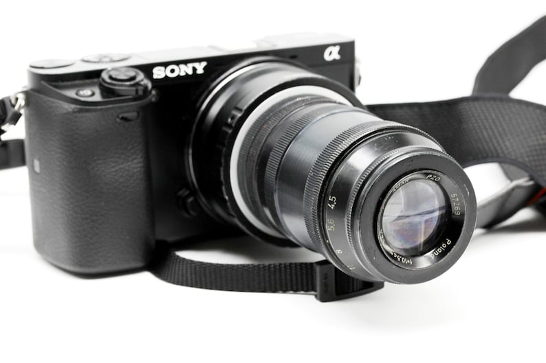 K&ouml;nnte beinhalten: Schwarze Sony Alpha Kamera mit gro&szlig;em Objektiv. Die Kamera hat einen schwarzen Riemen und befindet sich vor einem wei&szlig;en Hintergrund. Das Objektiv hat Messmarkierungen.
