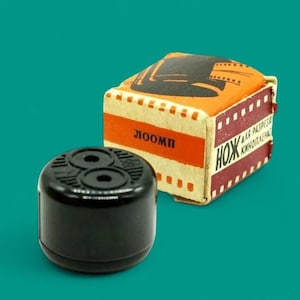 Puede incluir: Un objeto cilíndrico negro con dos aberturas circulares se encuentra frente a una pequeña caja rectangular. La caja es de color naranja y crema con detalles en rojo y texto en idioma extranjero. El fondo es turquesa.