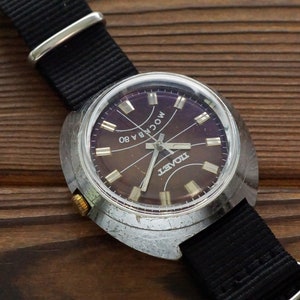 Pode incluir: Um rel&oacute;gio de pulso prateado com uma pulseira de couro preta. O mostrador do rel&oacute;gio &eacute; roxo escuro com ponteiros e marcadores prateados. O mostrador do rel&oacute;gio diz "Raketa" e "MOCKBA 80".