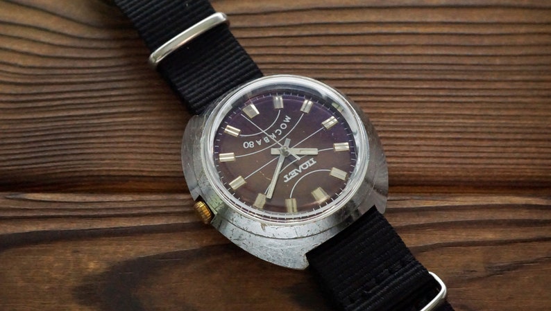 Pode incluir: Um rel&oacute;gio de pulso prateado com uma pulseira de couro preta. O mostrador do rel&oacute;gio &eacute; roxo escuro com ponteiros e marcadores prateados. O mostrador do rel&oacute;gio diz "Raketa" e "MOCKBA 80".