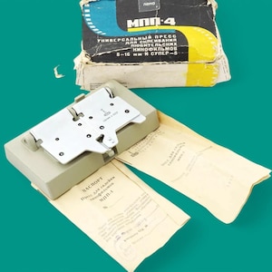 May include: A vintage film splicer kit with a gray base, a silver metal plate, and a box with Cyrillic text. The box is black, yellow, and blue. The kit includes paperwork with Cyrillic text. The background is a teal color.