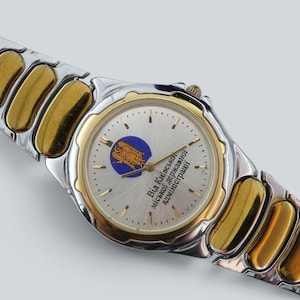 May include: A two-tone wristwatch featuring a silver and gold-colored metal link band. The round watch face has a silver background, a gold-colored bezel, and a blue circular emblem. The text "Від Київської міської державної адміністрації" is printed on the dial.