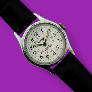 Könnte beinhalten: Eine silberne Armbanduhr mit weißem Zifferblatt und schwarzem Lederarmband. Das Zifferblatt der Uhr hat schwarze Zahlen und rote Markierungen. Das Zifferblatt der Uhr hat auch den Text "CARDINAL" und "21 JEWELS SHOCKPROOF".