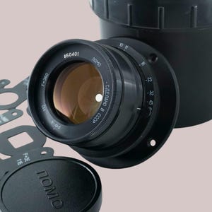May include: A black camera lens with the text "850401" and "F-360" is attached to a black cylindrical component. Several black aperture discs and a lens cap with the word "ПОМО" are also present. The lens has a gold-colored interior.