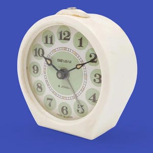 Pode incluir: Despertador vintage creme com mostrador verde claro e ponteiros pretos. O mostrador apresenta números pretos e a marca "SEVANI". O relógio está sobre um fundo azul.