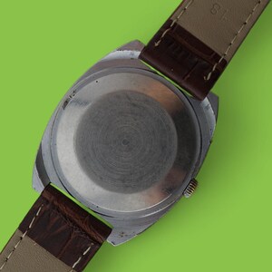 May include: Close-up of a vintage wristwatch with a silver-colored case and a brown leather strap. The watch back is visible, showing a circular design. The strap is marked "GENUINE LEATHER".