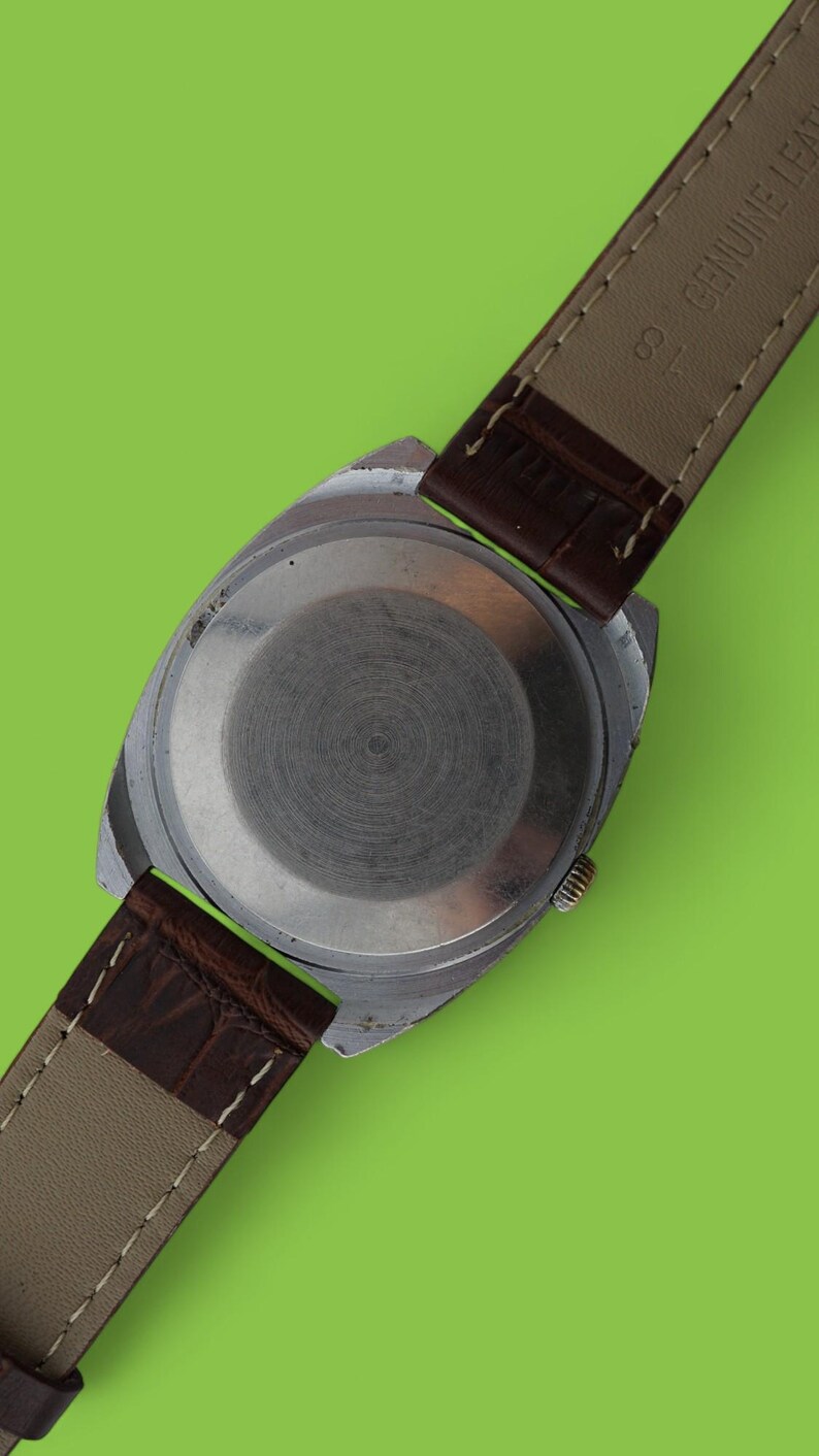 May include: Close-up of a vintage wristwatch with a silver-colored case and a brown leather strap. The watch back is visible, showing a circular design. The strap is marked "GENUINE LEATHER".