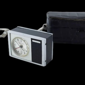 Pode incluir: Um despertador de viagem vintage com mostrador branco, caixa preta e prateada e estojo de couro preto. O relógio tem uma alça para pendurar e um pequeno alto-falante. O mostrador do relógio tem números e ponteiros pretos.