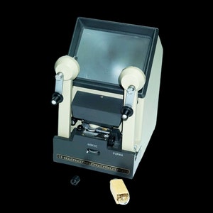 Puede incluir: Un proyector de películas vintage de color gris y beige. Tiene dos botones en el lateral y una pantalla en la parte delantera. El proyector tiene un botón de enfoque y un botón de cuadro etiquetados como "ФOКУС" y "РАМКА".