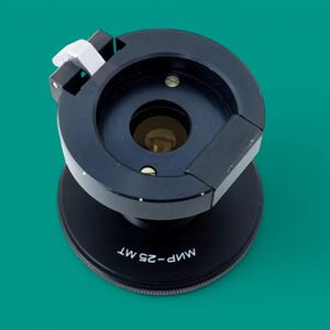 May include: A black camera lens adapter featuring a circular mount with a white locking lever and a central glass element. The base of the unit is marked with the white text "МИР-25 МТ" printed along the curved edge of the black metal housing.