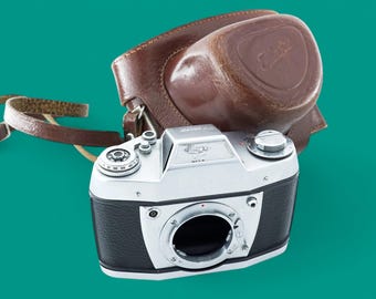 Puede incluir: Una cámara de película de 35 mm plateada y negra con una funda de cuero marrón. La cámara tiene un objetivo con un anillo negro alrededor y un dial plateado en el lateral.