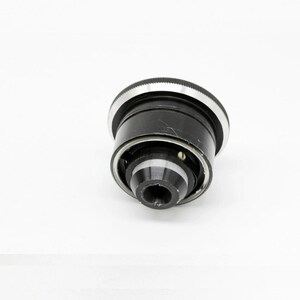 K&ouml;nnte beinhalten: Ein schwarzes Kameraobjektiv mit einem silbernen Ring oben. Das Objektiv hat konzentrische Ringe und eine zentrale Blende. Das Objektiv ist vor einem wei&szlig;en Hintergrund platziert.