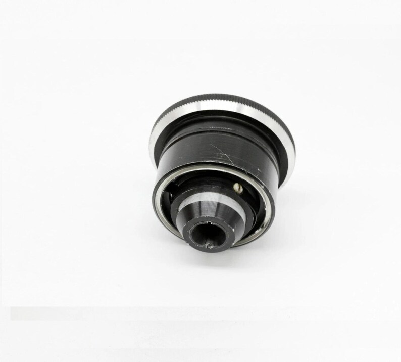 K&ouml;nnte beinhalten: Ein schwarzes Kameraobjektiv mit einem silbernen Ring oben. Das Objektiv hat konzentrische Ringe und eine zentrale Blende. Das Objektiv ist vor einem wei&szlig;en Hintergrund platziert.