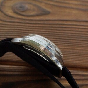 Pode incluir: Um rel&oacute;gio de pulso prateado com uma pulseira de nylon preta. O rel&oacute;gio est&aacute; sobre uma superf&iacute;cie de madeira.