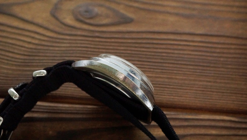 Pode incluir: Um rel&oacute;gio de pulso prateado com uma pulseira de nylon preta. O rel&oacute;gio est&aacute; sobre uma superf&iacute;cie de madeira.