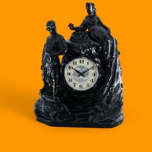 Puede incluir: Reloj negro con una esfera blanca y agujas negras. El reloj está rodeado por tres figuras negras, dos de pie y una sentada. El reloj está sobre una base negra.